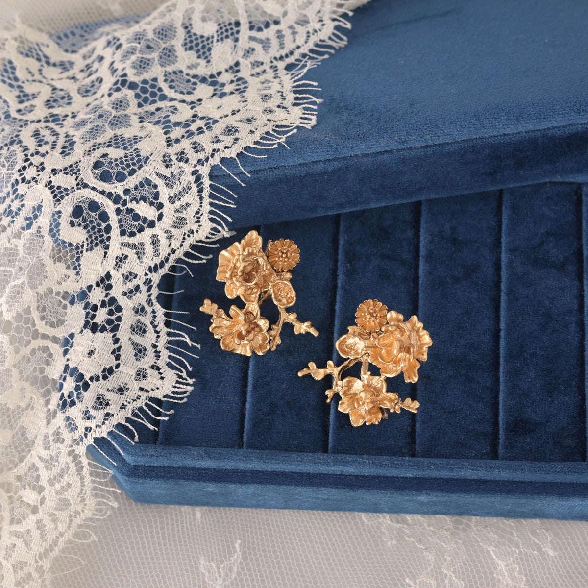A pair of gold floral stud earrings displayed on a blue velvet jewelry box, with white lace draped over the box on a white lace-covered surface.