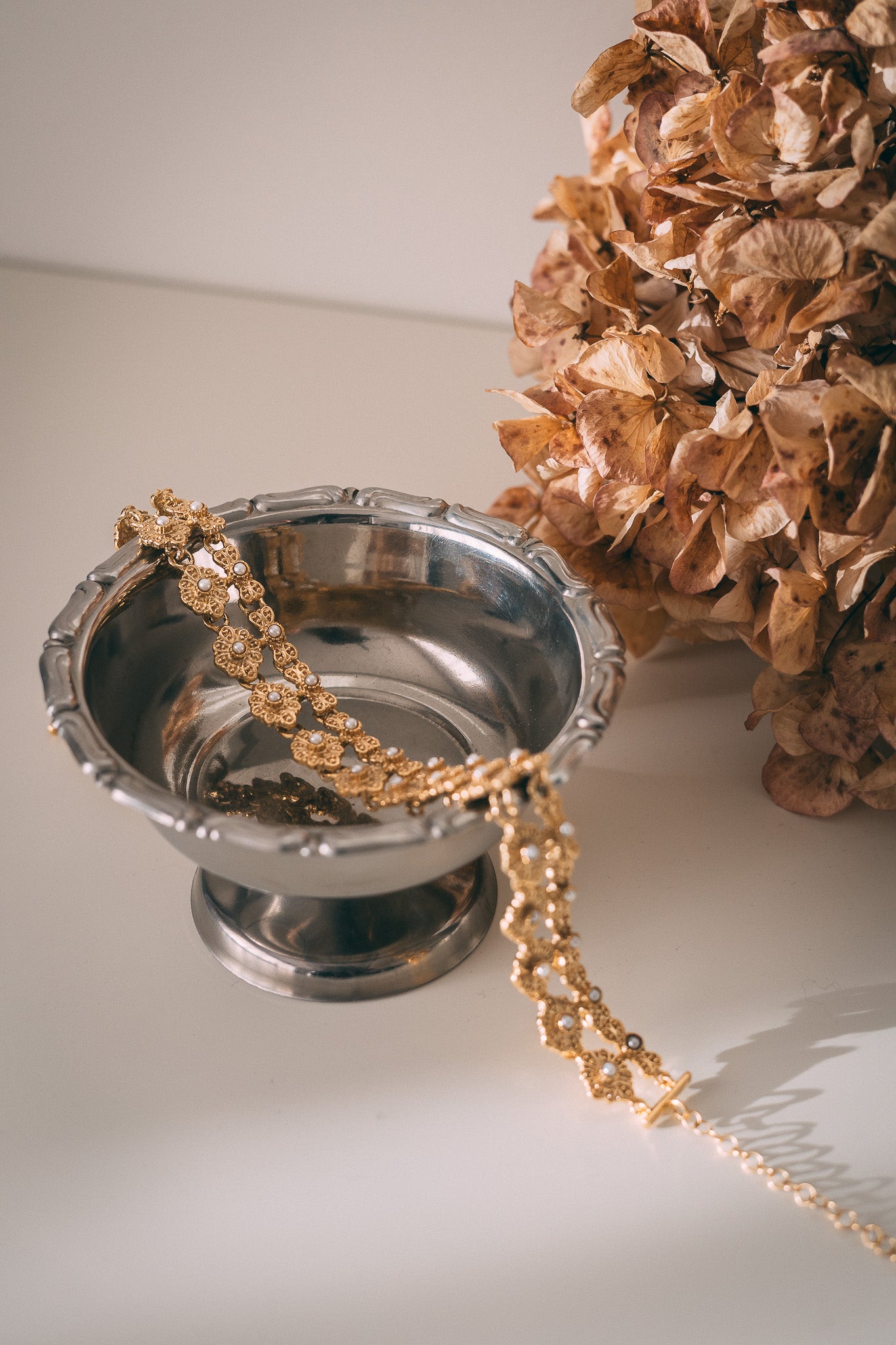 Silver bowl with a gold chain and dried flowers on a white surface