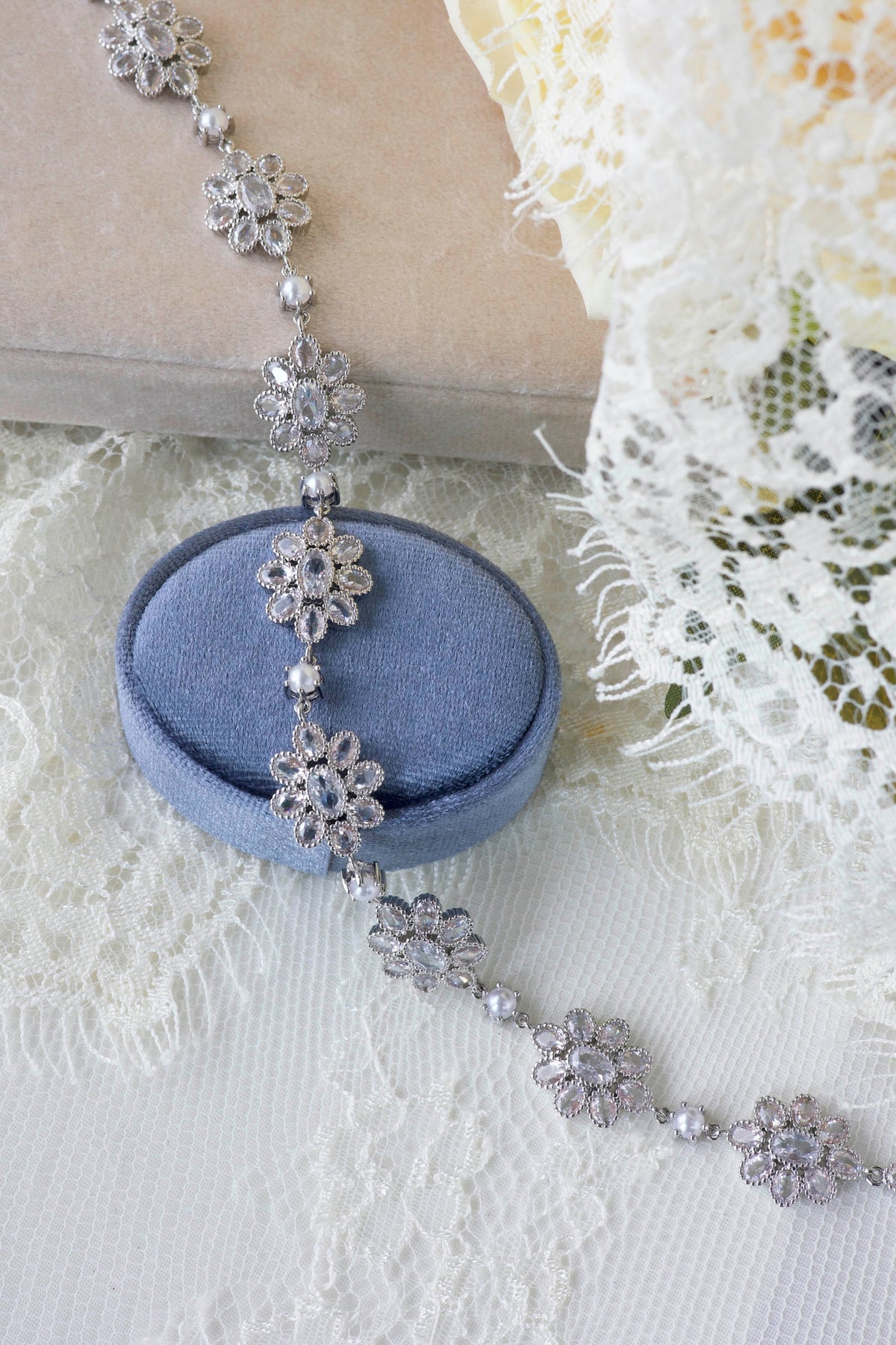 A silver-toned floral beaded necklace draped over a round blue velvet jewelry box, styled on white lace fabric with a beige book and pale yellow flower in the background.