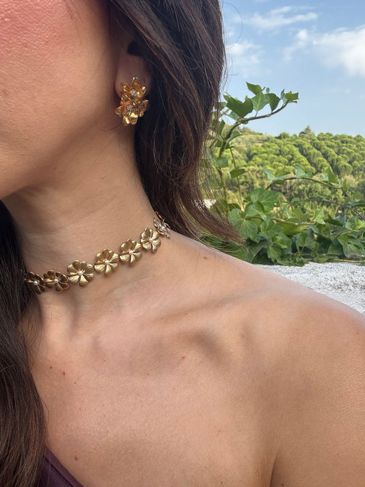 Close-up of a person wearing gold floral earrings and choker with a scenic background.