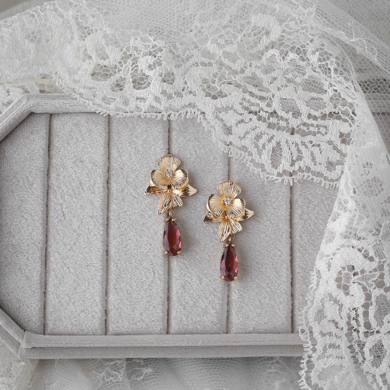 Gold floral earrings with garnet drops on a gray surface with lace fabric.
