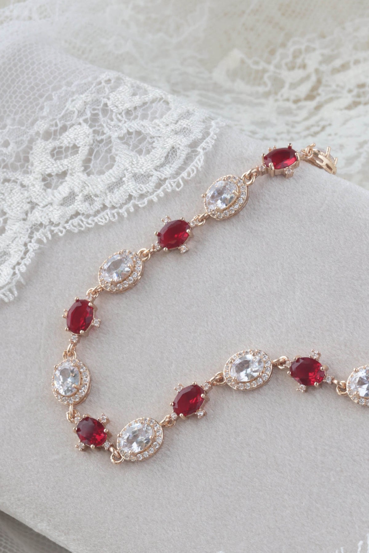 A rose gold chain bracelet with alternating oval red gemstones and clear oval gemstones, displayed on a soft white fabric with lace trim.