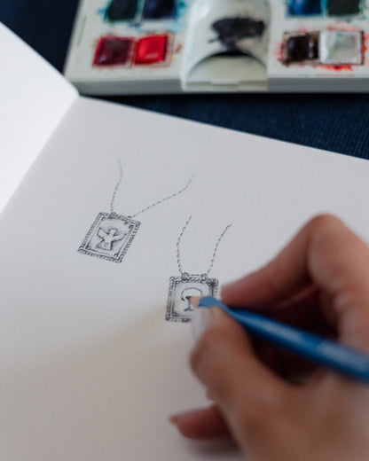 Hand drawing a necklace with a pendant on a sketchbook, with watercolor paints and brushes in the background.
