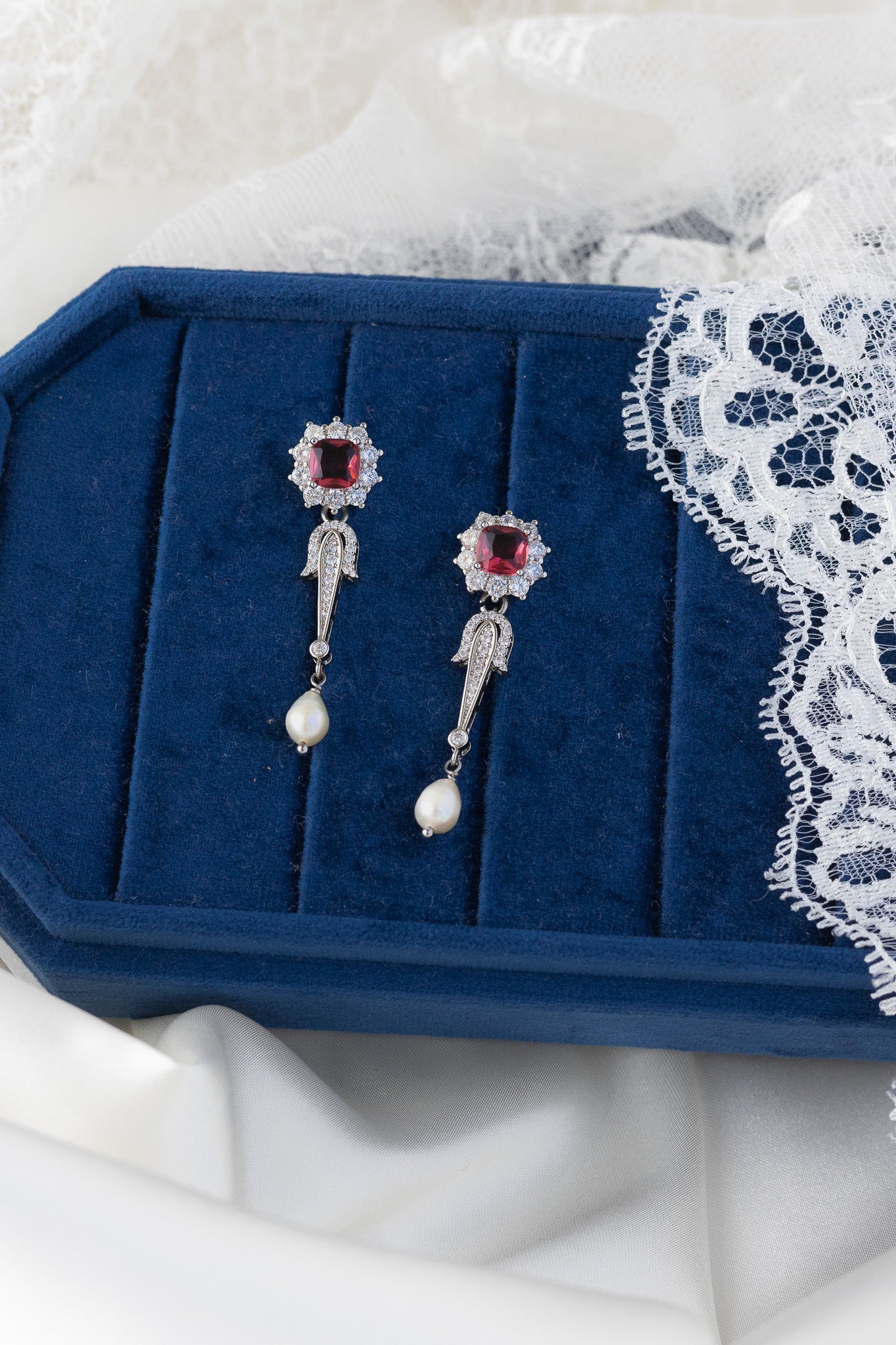 Pair of silver earrings with red gemstones on a blue jewelry box with lace trim.