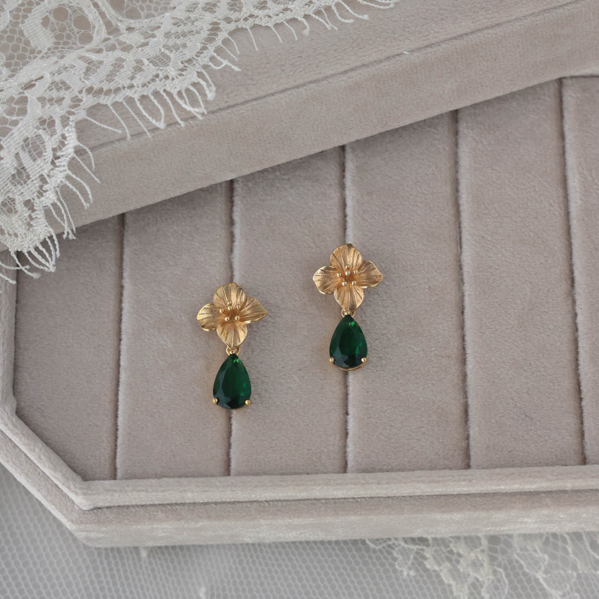 A pair of gold dangle earrings with flower-shaped tops and green teardrop gemstones, displayed on a beige jewelry box with a lace cloth underneath.