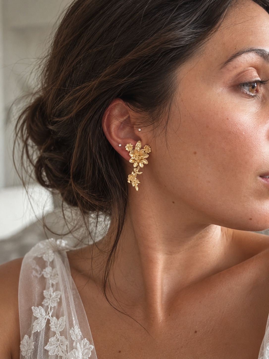 A close-up of a woman wearing a delicate gold floral drop earring, with her hair in an updo and a white lace-trimmed top visible.