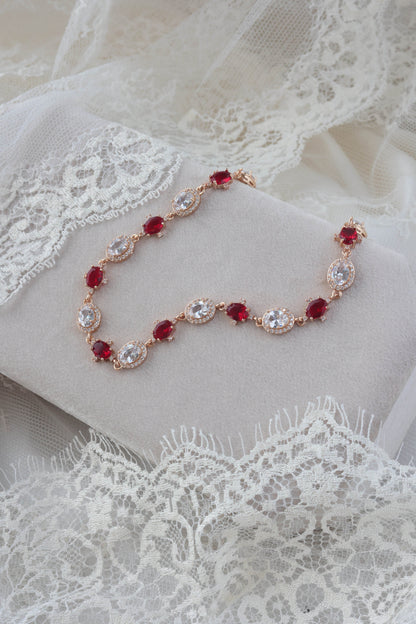 A rose gold chain bracelet with alternating oval red gemstones and oval clear gemstones, displayed on a white fabric surface with lace.