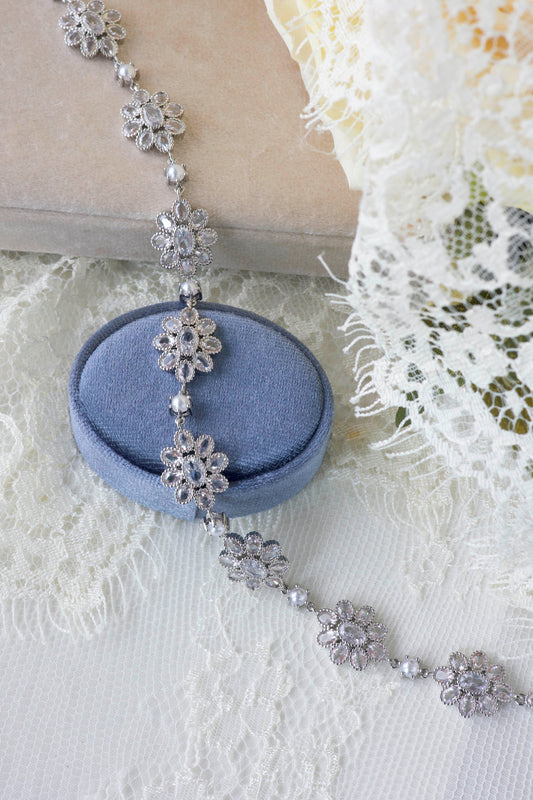 A silver-toned floral beaded necklace draped over a round blue velvet jewelry box, styled on white lace fabric with a beige book and pale yellow flower in the background.