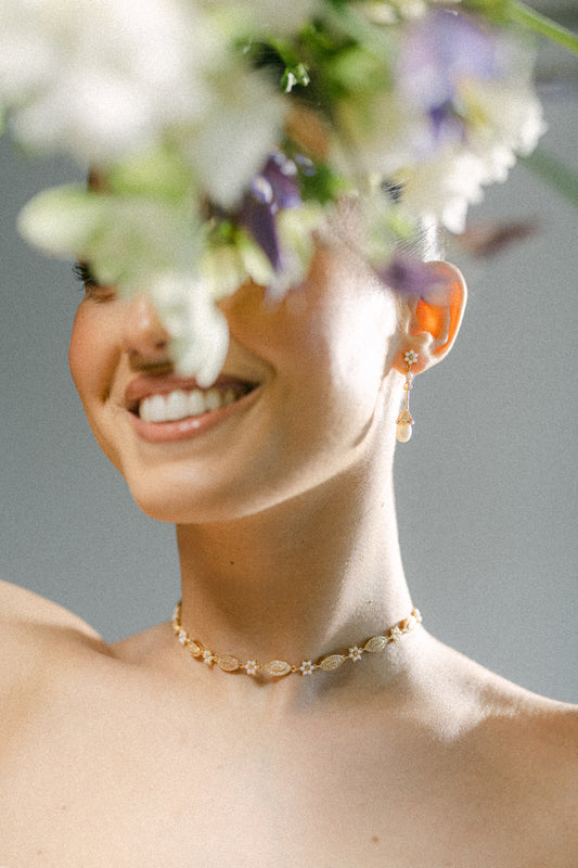 Woman with a gold necklace and earrings, smiling with flowers in the foreground.