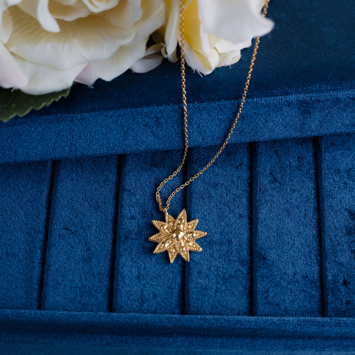 A golden starburst pendant necklace resting on a textured dark blue velvet jewelry box, with white roses in the upper left corner.