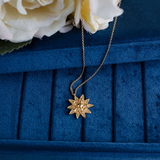 A golden starburst pendant necklace resting on a textured dark blue velvet jewelry box, with white roses in the upper left corner.